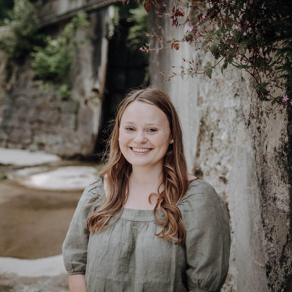 Eine lächelnde Frau mit braunem Haar trägt ein graues Kleid und steht vor einer Steinmauer mit Pflanzen im Hintergrund.