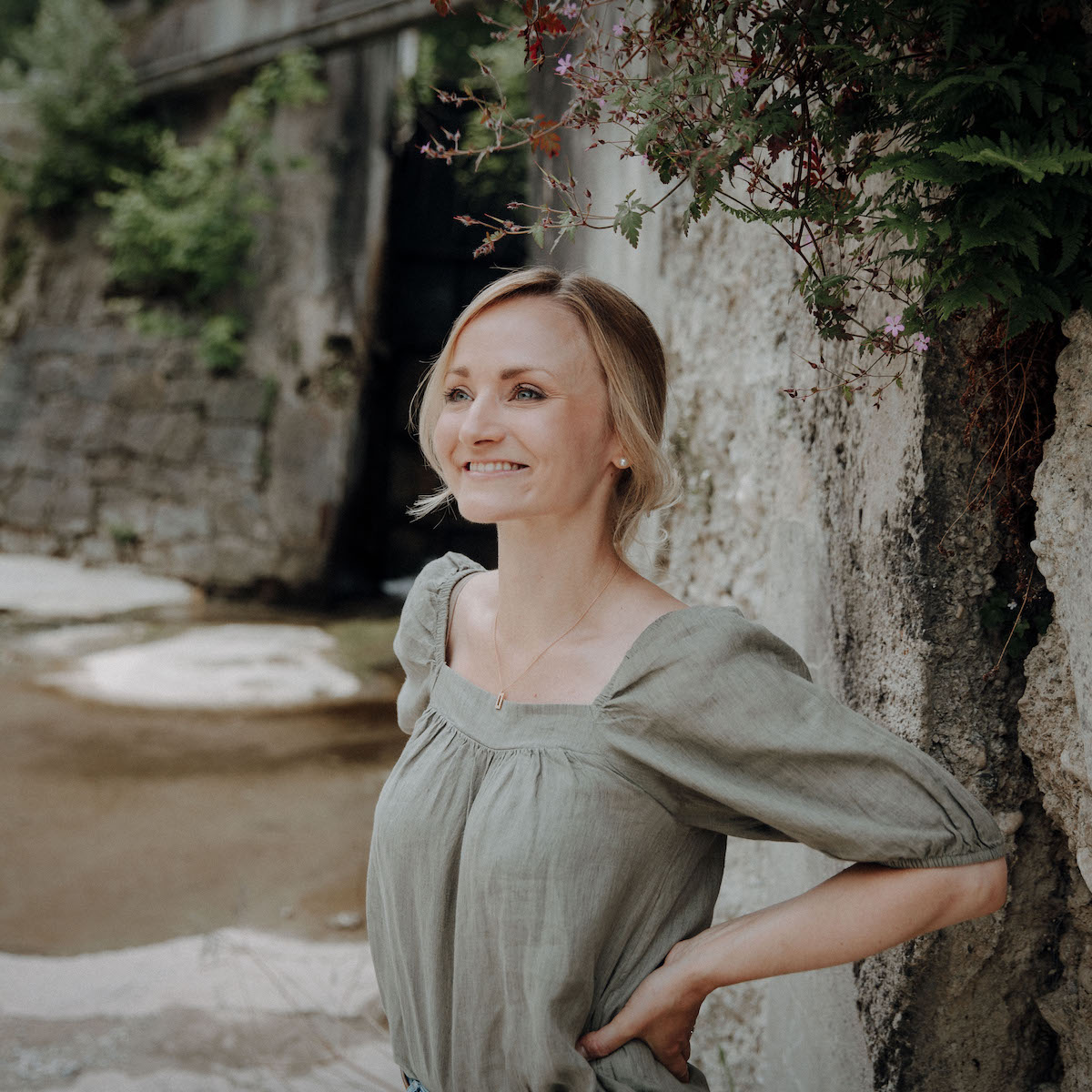 Eine lächelnde Frau in einem olivgrünen Kleid steht neben einer bewachsenen Steinmauer im Freien.