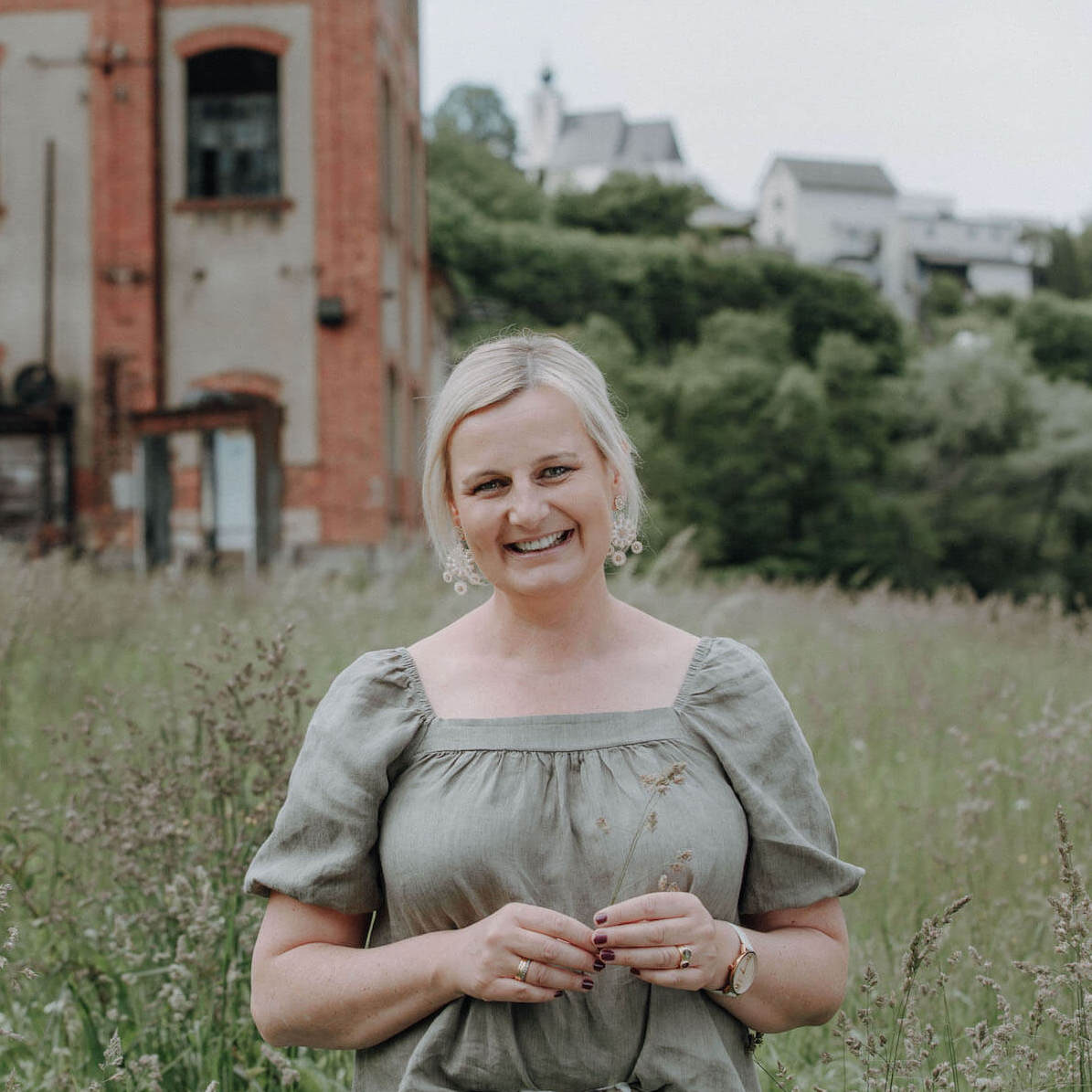 Lächelnde Frau in grünem Kleid steht auf einer Wiese vor einem alten Gebäude, im Hintergrund sind Bäume und weitere Häuser zu sehen.