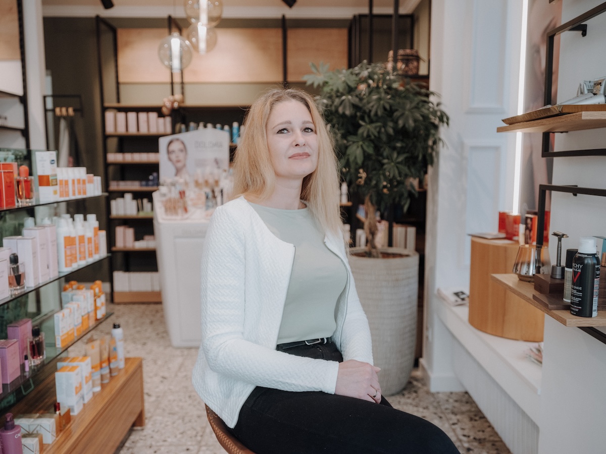 Eine Frau sitzt in einem Geschäft für Hautpflegeprodukte, umgeben von Regalen mit verschiedenen Kosmetikartikeln und Pflanzen.