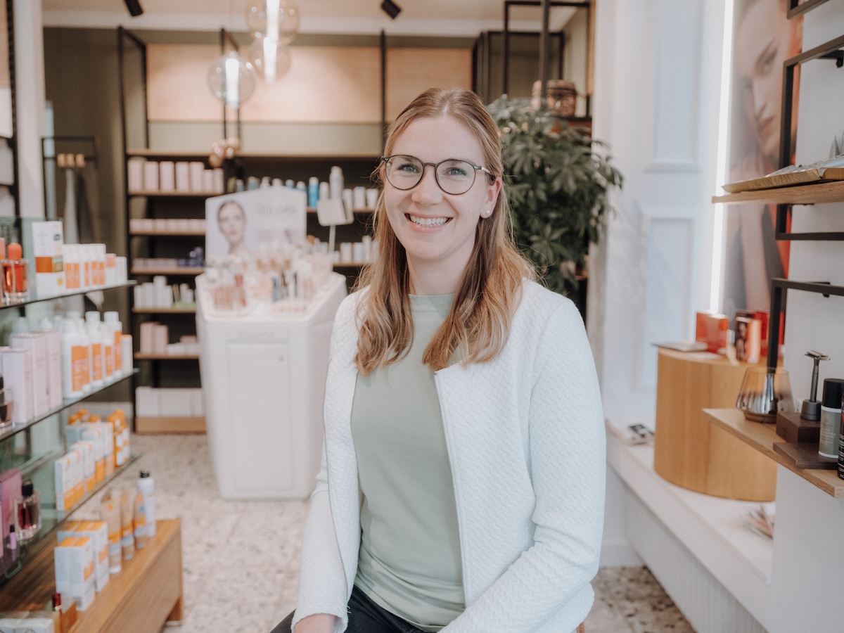 Eine lächelnde Frau mit Brille sitzt in einem Laden, umgeben von Kosmetikprodukten und Regalen, die mit Flaschen gefüllt sind.