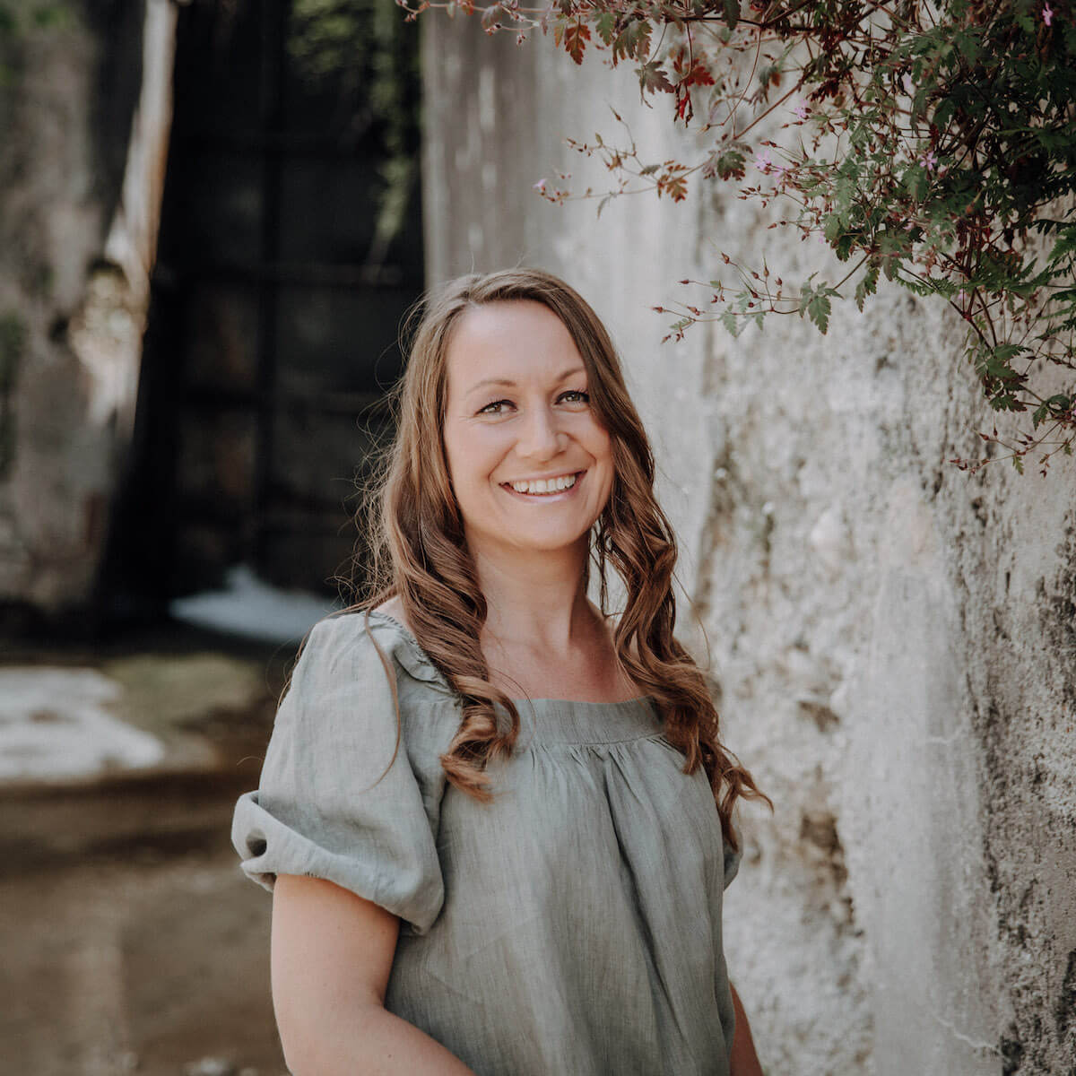 Eine lächelnde Frau mit langen, lockigen Haaren trägt ein graues Oberteil und steht neben einer Mauer mit blühenden Zweigen.