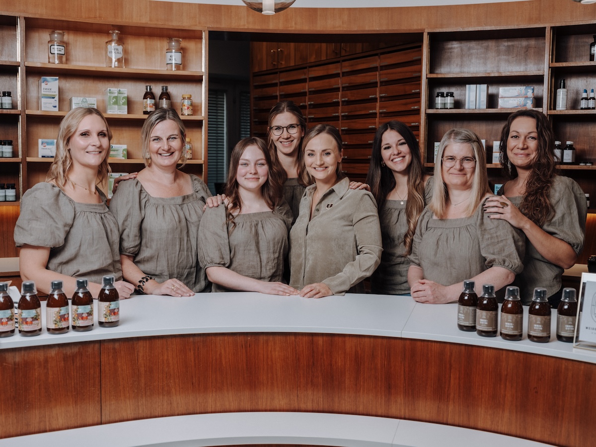 Eine Gruppe von acht Frauen steht lächelnd hinter einer Holztheke mit Getränkeflaschen, im Hintergrund befinden sich Regale mit Produkten.