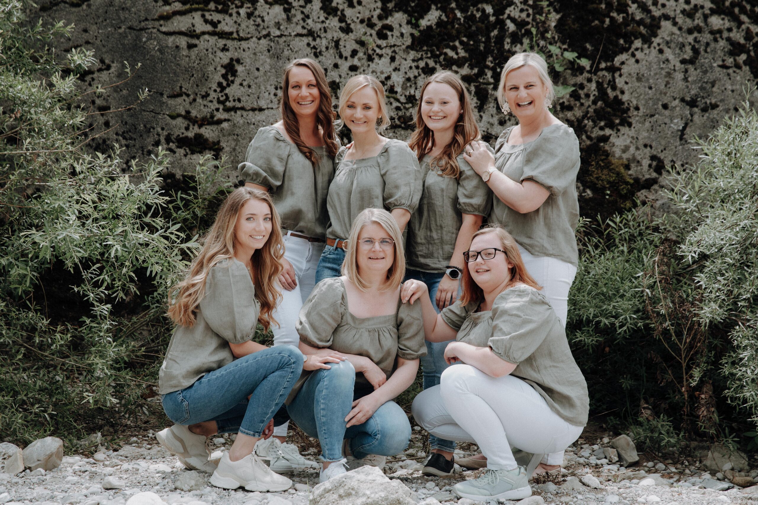 Sieben Frauen in ähnlicher Kleidung posieren lächelnd in der Natur vor einem Stein und Sträuchern.