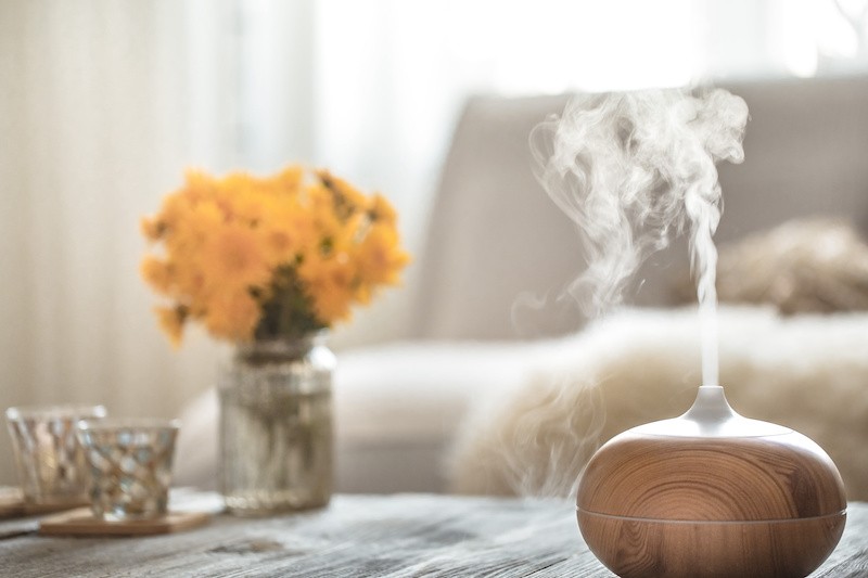 Ein Holz-Aromadiffuser verströmt Dampf auf einem Tisch, neben einer Vase mit gelben Blumen und zwei Teelichthaltern.