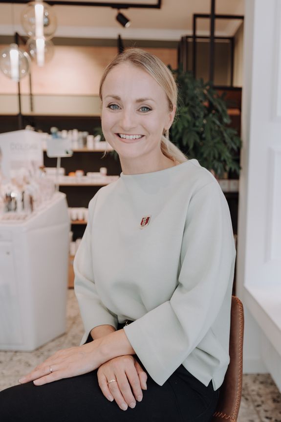 Eine lächelnde Frau sitzt in einem Geschäft mit Kosmetikprodukten, sie trägt einen hellen Pullover mit einer kleinen Anstecknadel.